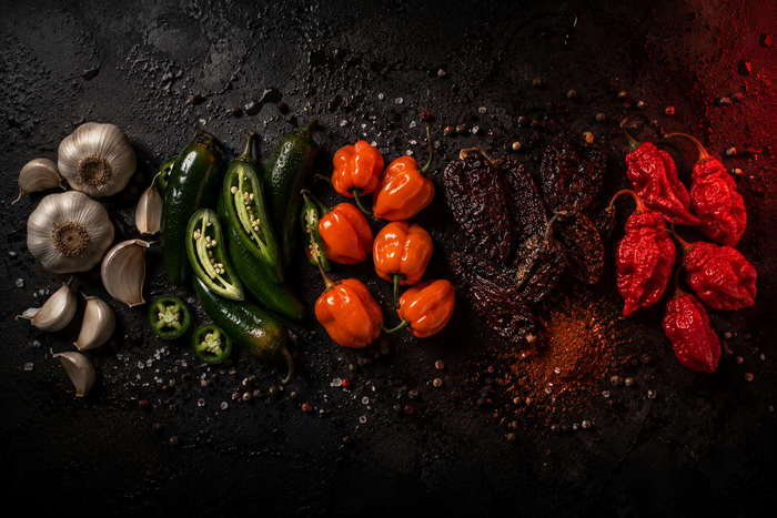 The five Bad Decision ingredients laid out left to right in heat order on dark stone: garlic bulbs, fresh jalapenos, orange habaneros, dried chipotles with ground chipotle, and red ghost peppers