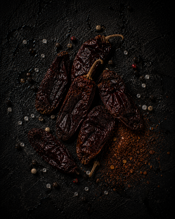 Dried smoked chipotle peppers, deep red-brown and wrinkled, on dark stone with a scattering of ground chipotle