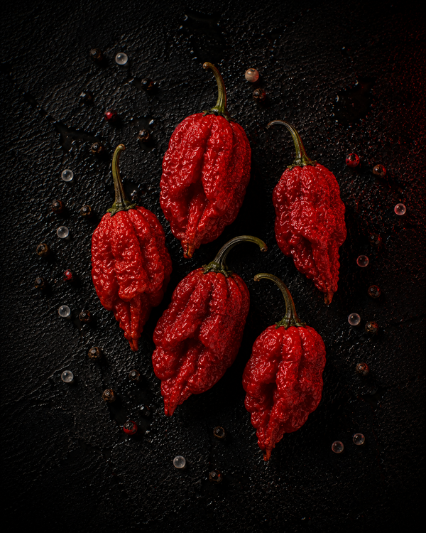 Five bright red ghost jolokia peppers with green stems on dark stone with scattered peppercorns