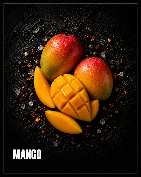 Two ripe mangoes and a scored mango half with slices, on dark stone with scattered chilli flakes and salt