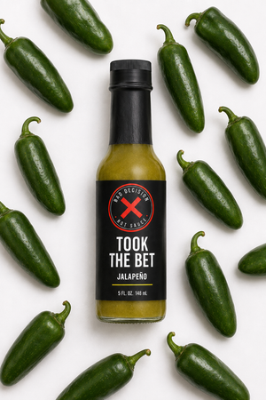 Took The Bet jalapeño hot sauce bottle lying flat among fresh jalapeños on a white background