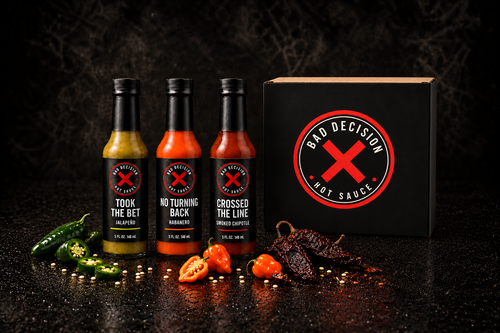 Bad Idea 3-Bottle Set — Took The Bet jalapeño, No Turning Back habanero and Crossed The Line smoked chipotle beside the black Bad Decision gift box, with fresh and dried chillies