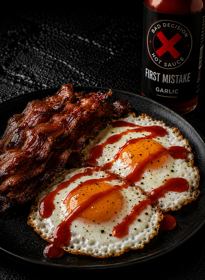 Two fried eggs and bacon drizzled with red garlic hot sauce, beside a bottle of First Mistake garlic hot sauce