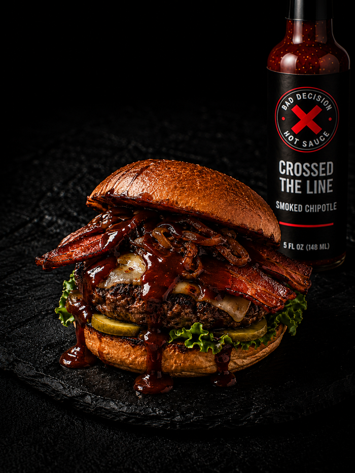 A bacon burger dripping with dark chipotle sauce, beside a bottle of Crossed The Line smoked chipotle hot sauce
