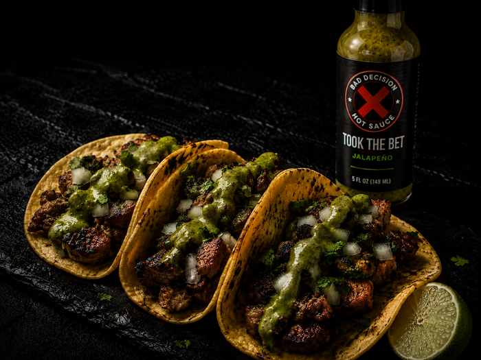 Three street tacos topped with green jalapeno sauce and onion, beside a bottle of Took The Bet jalapeno hot sauce