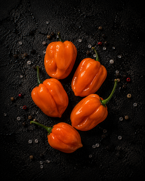 Five bright orange habanero peppers with green stems on dark stone with scattered peppercorns and salt