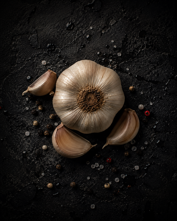 A whole garlic bulb and loose cloves on dark stone with scattered peppercorns and salt