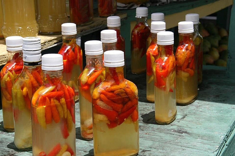 Bottles of Puerto Rican pique lined up on a weathered green table, whole red and orange hot peppers steeping in vinegar